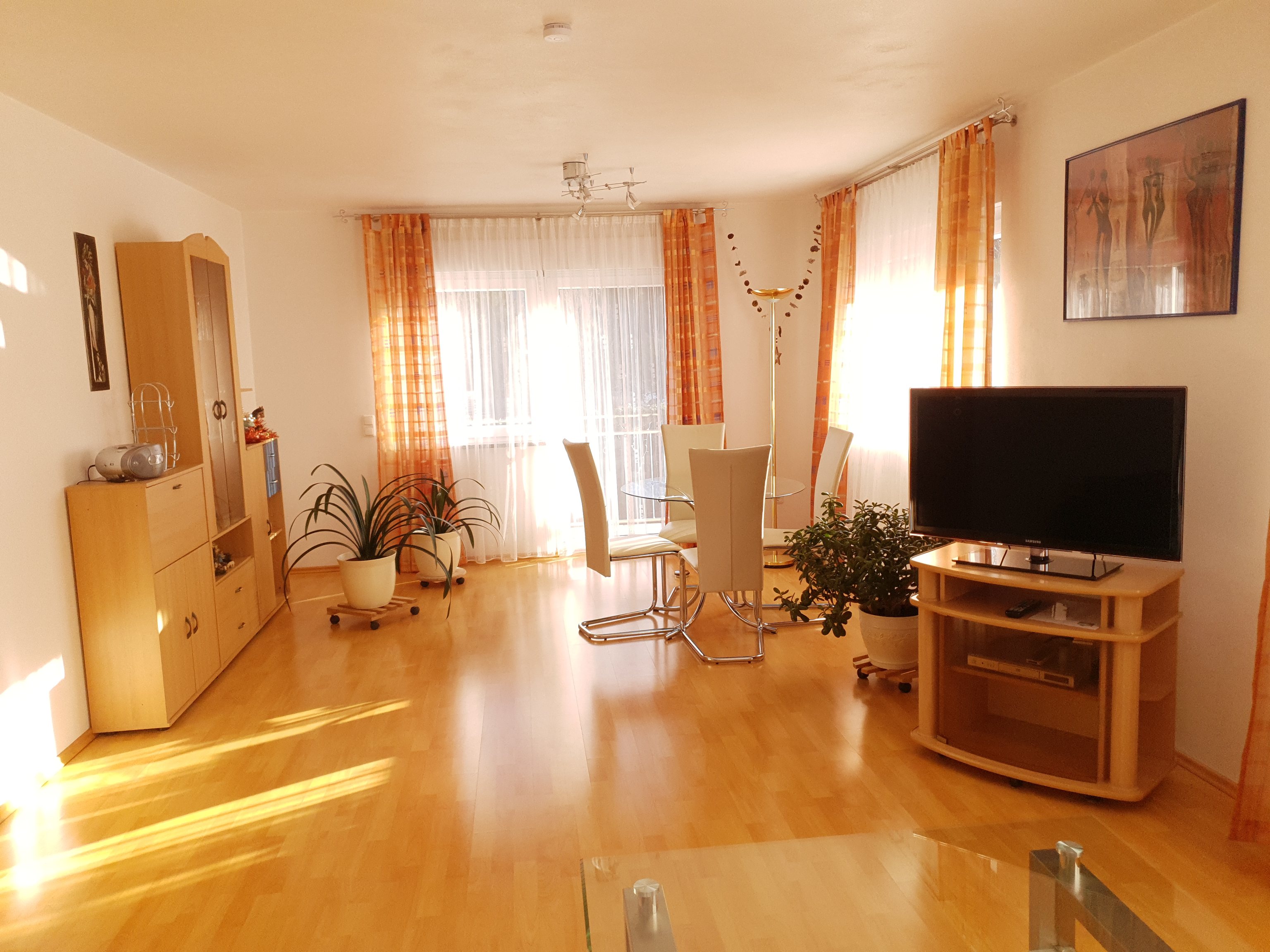 Wohn- und Essbereich mit hellen Möbeln, Esstisch aus Glas, Pflanzen und zwei Fenstern zum Balkon (Foto der Familie Bergmann, Januar 2024)