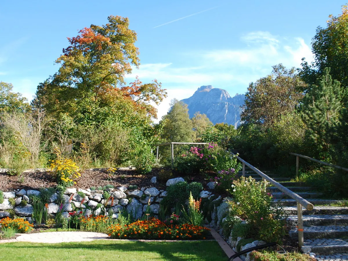 Garten mit Steinmauer, Holzstufen und Blumenbeeten im Herbst, dahinter ein Berg